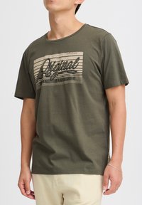 Tricou din bumbac de culoare verde măsliniu, cu guler rotund, mâneci scurte și un imprimeu texturat care prezintă cuvântul "Original" în negru și bej.