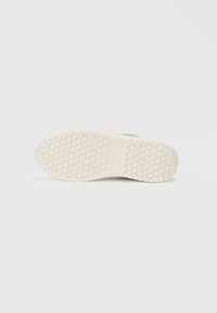 Semelle en caoutchouc avec un motif texturé de petits nubs circulaires. La couleur est blanc cassé avec un détail de marque légèrement en relief. Design plat et large.