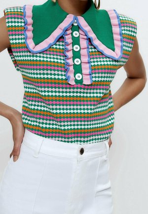 Ærmeløs strikbluse i grøn, pink, orange og hvid med diamantmønster, stor grøn krave, pink og blå flæser, hvide knapper, båret med hvide bukser.