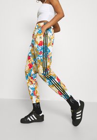 Färgglada joggers med blommönster i vitt, blått och gult, med svarta vertikala ränder och elastisk midja. Svarta sportskor med vita detaljer.