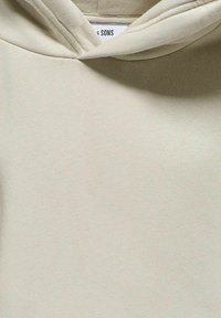 Ljusbeige hoodie med en texturerad, mjuk tyg. Har en dragsnodd i huvan och ett sydd märke vid nacken. Enkel, minimalistisk design.