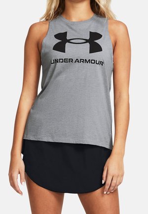Kobieta ubrana w szary top bez rękawów z czarnym logo Under Armour i czarne szorty sportowe, stojąca na jednolitym tle.