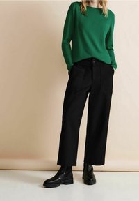 Grüner, langärmeliger Pullover mit Rundhalsausschnitt, kombiniert mit hochgeschnittenen schwarzen weit geschnittenen Hosen. Schwarze Knöchelstiefel runden das Outfit ab.