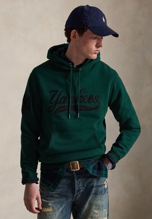 Jeune homme portant un sweat à capuche Yankees vert foncé, une casquette de baseball bleue, une chemise à carreaux, un jean usé et une montre-bracelet, regardant sur le côté.