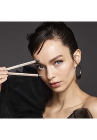 Deux crayons à sourcils beiges avec des accents argentés, tenus près du visage d'un modèle, avec un tissu noir transparent drapé sur son épaule. Un éclairage doux met en valeur ses traits.