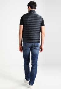 Black puffer vest with vertical stitching, high neck, and zip closure; paired with blue jeans and white sneakers, shown from the back.