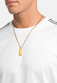 Collana in oro con pendente a forma di tag rettangolare, indossata sopra una camicia bianca semplice con dettagli neri sulle spalle.
