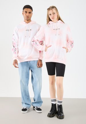 Lyserøde tie-dye hættetrøjer har blomsterbroderi på ærmerne. Den mandlige model har lyseblå jeans og sorte sneakers på; den kvindelige model bærer sorte shorts og støvler.