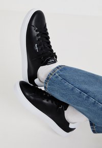 Czarne skórzane sneakersy z białą podeszwą, charakteryzujące się gładką fakturą, sznurowanym zapięciem i logo na boku. Dżinsy z detalem mankietu.