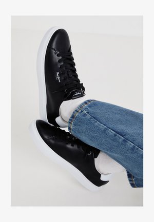 Fekete bőr sportcipők fehér talppal, sima textúrával, fűzős kialakítással és oldalsó logóval. Farmer, amelynek részlete a szegés.