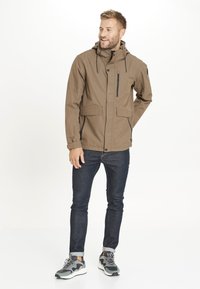Brun vattentät jacka med en dragkedjeförsedd bröstficka, justerbar huva och två framfickor. Bärs med mörka jeans och grå sneakers.