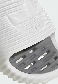 Vit slip-on sandal med ribbad textur och logotypdetalj, med en grå, ventilerad sula med utskärningar för andningsförmåga.