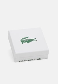Baltas stačiakampis dėžutė su tekstūruotu paviršiumi, kurioje yra žalias krokodilo logotipas ir žodžiai "LACOSTE" atspausdinti žalia spalva šone.