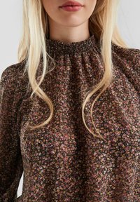 Vrouw met lang blond haar draagt een blouse met hoge kraag, versierd met een kleine bloemenprint in bruine, gele en roze tinten.