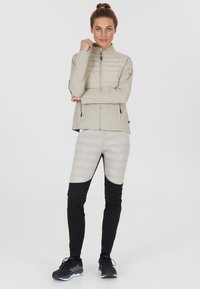 Lätt beige zip-up jacka med quiltad front, kombinerad med grå och svarta tajta byxor med en mjuk yta. Svarta träningsskor.