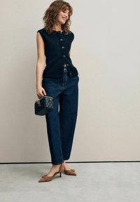 Top senza maniche in maglia nera con chiusura a bottoni, jeans blu a vita alta, mule con tacco a punta marroni e una piccola borsa a tracolla nera trapuntata.