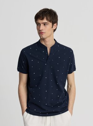 Giovane uomo che indossa una maglietta Henley blu navy a maniche corte con piccoli motivi di stelle bianche, in piedi con le mani in tasca, che guarda verso sinistra.