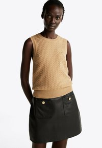 Beige kabelgebreide mouwloze trui met een ronde halslijn, gecombineerd met een zwarte leren minirok met gouden knopen en voorzakken.