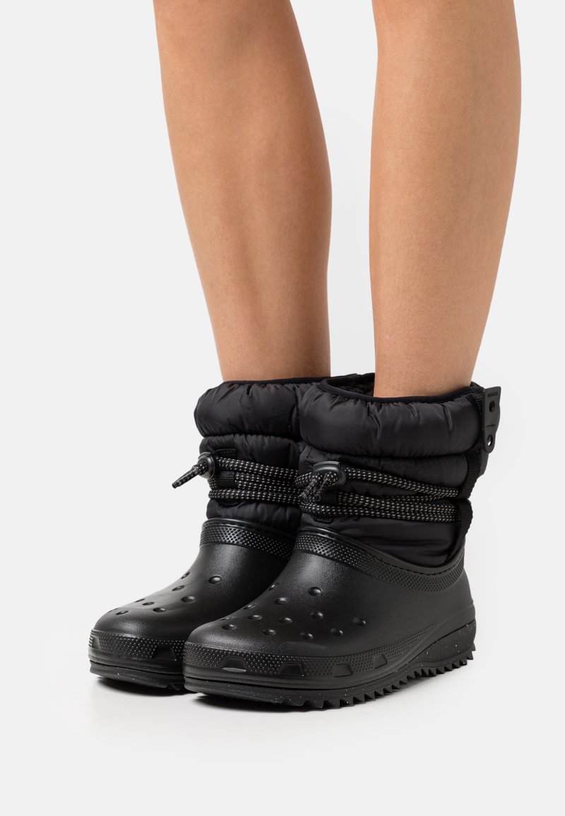 Bottes noires imperméables avec un dessus rembourré, fermeture par cordon de serrage et semelle texturée. Le design comprend des trous de ventilation et une forme massive.