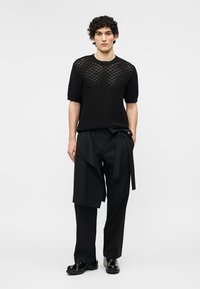 Άνδρας που φοράει μαύρο πλεκτό πουλόβερ με κοντά μανίκια, μαύρο φαρδύ παντελόνι με λεπτομέρεια από ύφασμα και ζώνη, και μαύρα γυαλιστερά παπούτσια, όρθιος μπροστά σε λευκό φόντο.