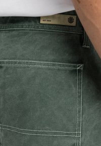 Pantalons en coton vert foncé avec une finition texturée, dotés d'une poche arrière et d'une étiquette portant la mention "EST. 1992". Coutures blanches en accent.