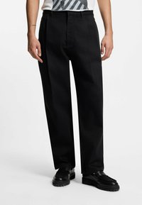 Pantaloni neri a gamba larga con una texture liscia e pieghe anteriori, abbinati a scarpe nere. La vita presenta passanti per una cintura standard.