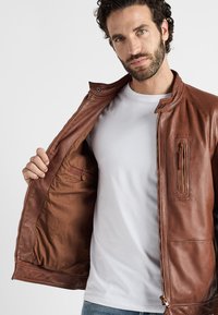 Braune Lederjacke mit Reißverschluss, ausgestattet mit seitlichen und Brusttaschen sowie einer glatten, matten Oberfläche. Futter aus braunem Stoff.