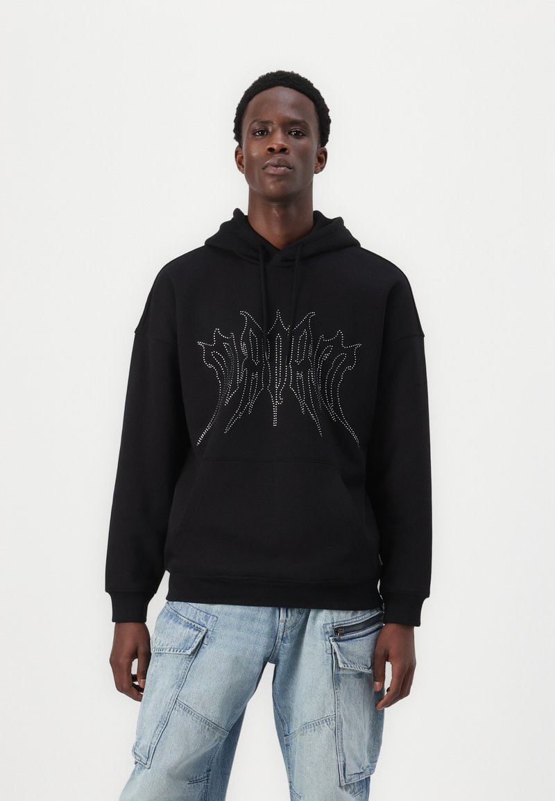 Black hoodie with a front graphic in silver studs, featuring a drawstring hood and ribbed cuffs. Paired with light blue cargo jeans.