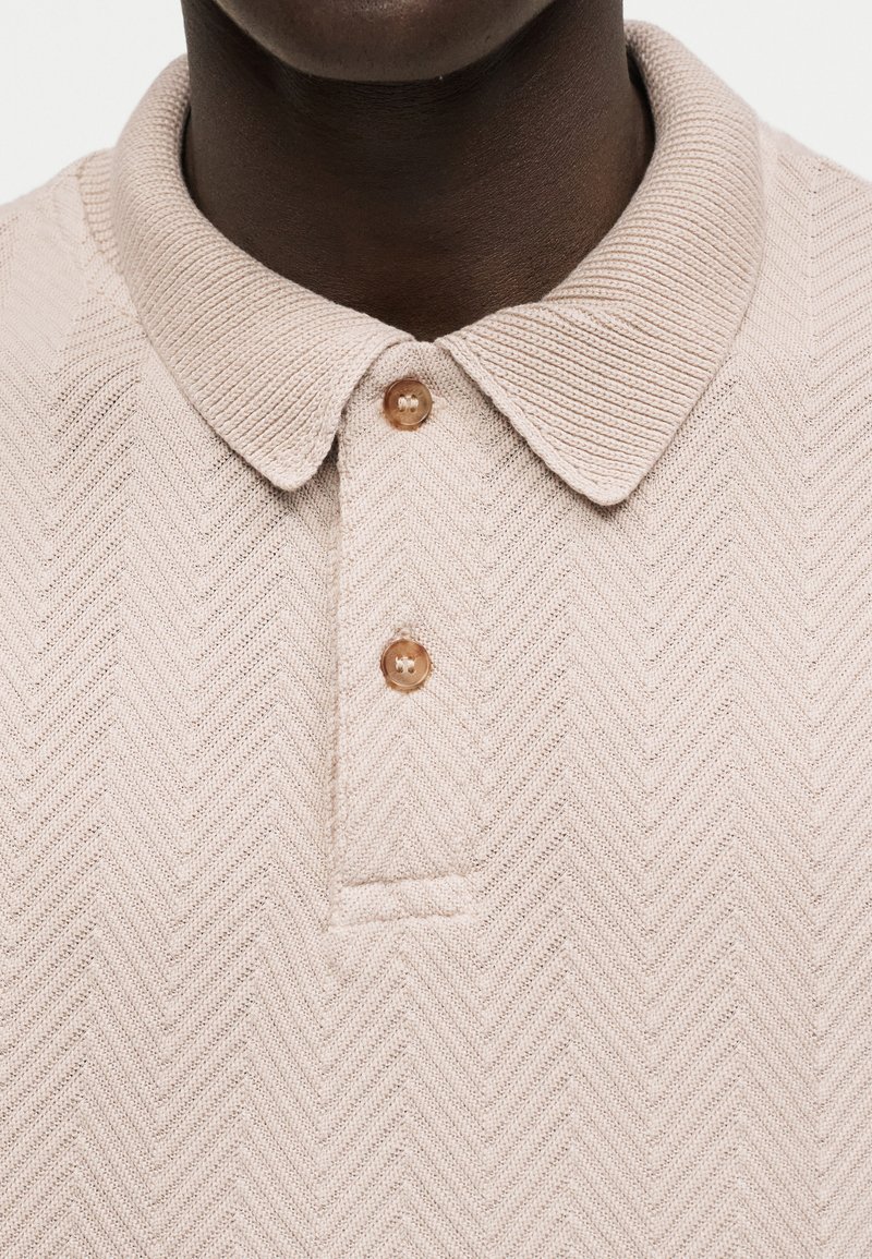 Gros plan d'une personne à la peau foncée portant un polo en maille beige avec un motif chevron et deux boutons marron fermés au col.