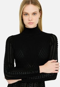 Haut en tricot noir avec un col montant, présentant une texture en côtes verticales et des motifs découpés. Les manches longues arborent des accents tressés sur les côtés.