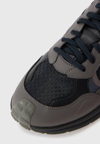 Gray sneaker featuring textured mesh and smooth leather panels, dark blue laces, and a rubber outsole with a patterned tread.