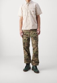Korte mouwen beige overhemd met knopen en een borstzak, gecombineerd met camouflage cargobroek en donkergroene veterschoenen.