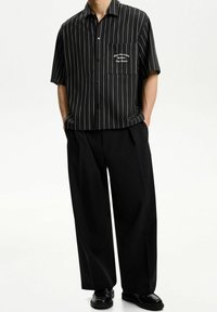 Černé tričko s krátkým rukávem, bílé vertikální pruhy a vyšívaná kapsa, spojené s volnými černými kalhotami a černými botami.