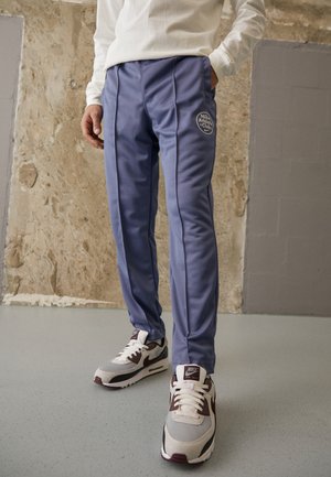 Light blue athletic pants with logo on left thigh, featuring a smooth, stretchy fabric and contrasting side seams. Paired with sneakers.