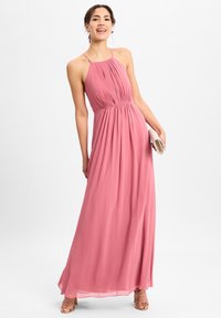 Maxi-jurk in zachtroze chiffon, met een geplooide lijfje, halternek en een vloeiende rok. Model houdt een kleine clutch vast en draagt sleehakken.