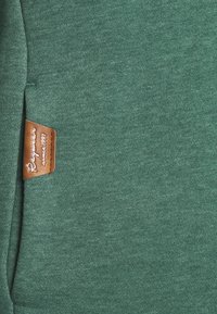 Felpa verde realizzata in un tessuto morbido, con un'etichetta in pelle su cui è ricamato in bianco "Raquetar dal 1997". Texture liscia, senza motivi.