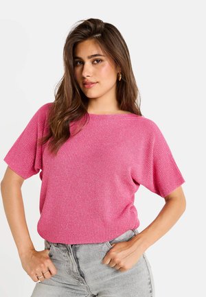Pull en tricot rose à manches courtes avec un motif côtelé texturé et une coupe décontractée, porté avec un jean taille haute gris, les mains sur les hanches.