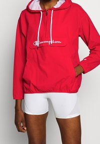 Person som bär en röd Champion-hoodie med vita snoddar och vita tighta shorts, står med en hand i fickan.