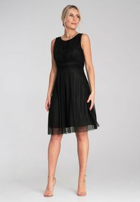 Schwarzes ärmelloses Kleid mit einem plissierten Oberteil und tailliertem Bund; fließt in einen knielangen Rock mit transparenter Überlage. Schlichtes Design und klassischer Ausschnitt.