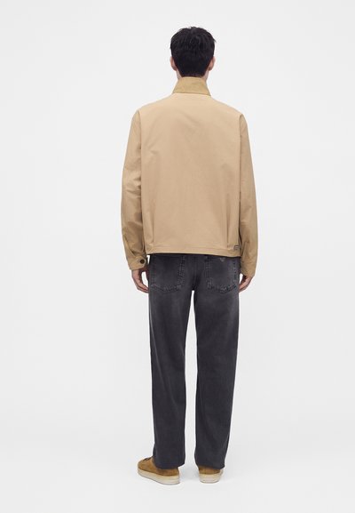 Homme debout de dos portant une veste beige, un jean gris foncé et des baskets marron sur un fond blanc uni.