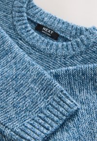 Kék kötött pulóver bordázott nyakkal és mandzsettákkal, különböző kék árnyalatokból álló mintázattal. Címke látható a nyaknál.