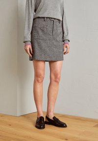 Grau karierter Rock mit zwei Vorderentaschen, Gürtelschlaufen und geradem Schnitt. Getragen mit dunkelbraunen Slippers und einem grauen Langarmoberteil.