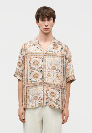 Νεαρός άντρας που φοράει κοντομάνικο πουκάμισο με κουμπιά, με σχέδιο λουλουδιών και ήλιου σε μπεζ, πορτοκαλί και πράσινους τόνους.