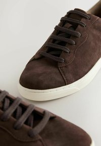 Baskets en daim marron avec des lacets marron foncé et des semelles en caoutchouc blanches, présentées sur une surface gris clair.