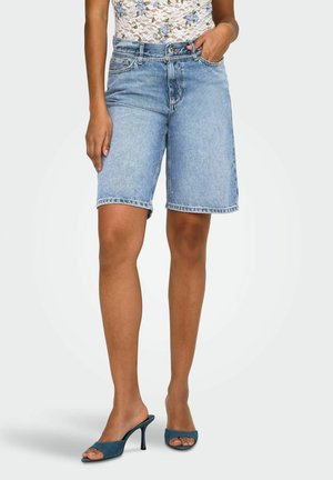 Žena oblečená v svetlomodrých džínsových šortkách po kolená s bielym kvetinovým čipkovaným topom a tyrkysovými sandálmi na podpätku s otvorenou špičkou, stojaca s jednou rukou v vrecku.