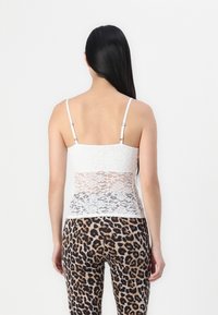 Camisole en dentelle blanche avec des bretelles ajustables, ornée de motifs floraux, superposée à un tissu blanc uni. Portée avec des leggings à motif animal.