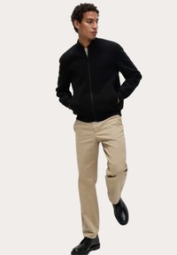 Zwarte bomberjack met rits aan de voorkant, geribde manchetten en zakken, gecombineerd met beige broeken en zwarte schoenen, op een lichte achtergrond.