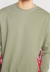 Sudadera verde oliva con mangas largas, cuello redondo acanalado y acentos laterales rojos con texto blanco. Tejido suave y texturizado.