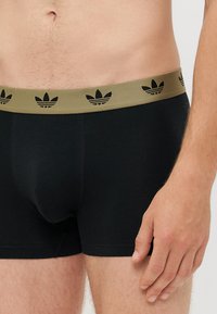 Črne bokserice z širokim olivno-zelenim pasom, ki ima črn vzorec Adidas s tremi črtami. Mehka, prilegajoča se zasnova za udobje.