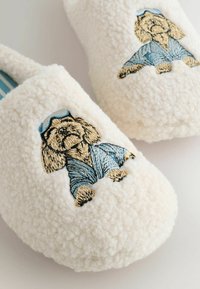 Krämmfärgade tofflor gjorda av fluffigt material, med broderad hunddesign i blå kläder och randig innerfoder.
